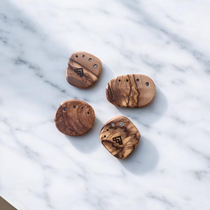 May include: Four olive wood salt and pepper cellars with a natural wood grain pattern. Each cellar has a unique shape and features small holes for dispensing. A black logo is visible on each cellar.