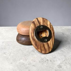 May include: Wooden bottle opener with a brass opener, featuring a natural wood grain pattern. The opener is oval-shaped and rests on top of two wooden coasters in varying shades of brown.