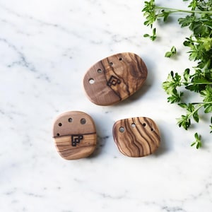 May include: Three olive wood soap dishes with a natural wood grain pattern. Each dish has a unique shape and several drainage holes. A small, dark logo is etched into each dish. The dishes are arranged on a white marble surface.