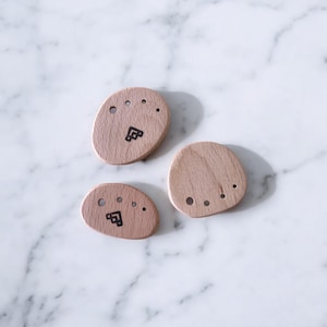 May include: Three wooden, oval-shaped objects with a light brown finish. Each has a small, black graphic and several small holes. The objects are arranged on a white marble surface, creating a minimalist aesthetic.