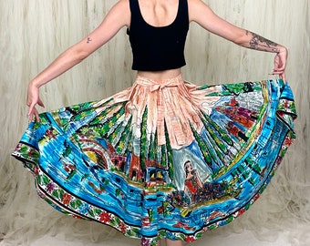 May include: A full-length view of a woman wearing a colorful, printed skirt with a scene of a village and a woman in a boat. The skirt is gathered at the waist and has a wide, flowing silhouette. The woman is wearing a black tank top and has her hair pulled back in a bun.