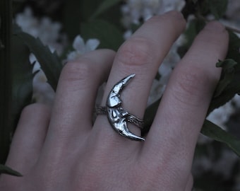 May include: A silver crescent moon ring with a textured surface. The ring is worn on a person's finger.