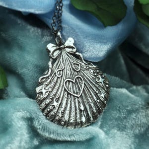 May include: A silver-toned pendant necklace featuring a seashell design with a heart and star accents. The pendant is suspended from a delicate chain and is set against a backdrop of blue fabric and green foliage. The pendant has a bow at the top.
