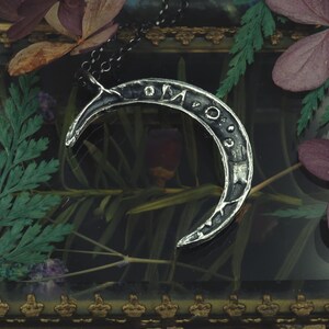 May include: A silver crescent moon pendant necklace with an inscription on the surface. The necklace is displayed in a glass box with dried flowers and greenery.