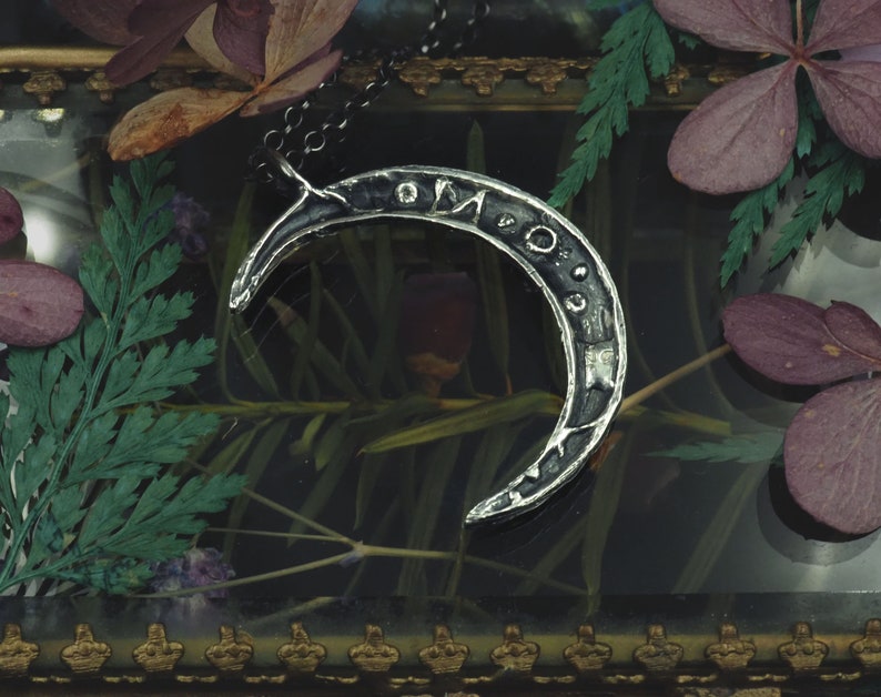 May include: A silver crescent moon pendant necklace with an inscription on the surface. The necklace is displayed in a glass box with dried flowers and greenery.