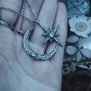 May include: A silver necklace with a crescent moon and star pendant. The pendant is detailed with intricate floral designs and features a clear gemstone in the center of the star.
