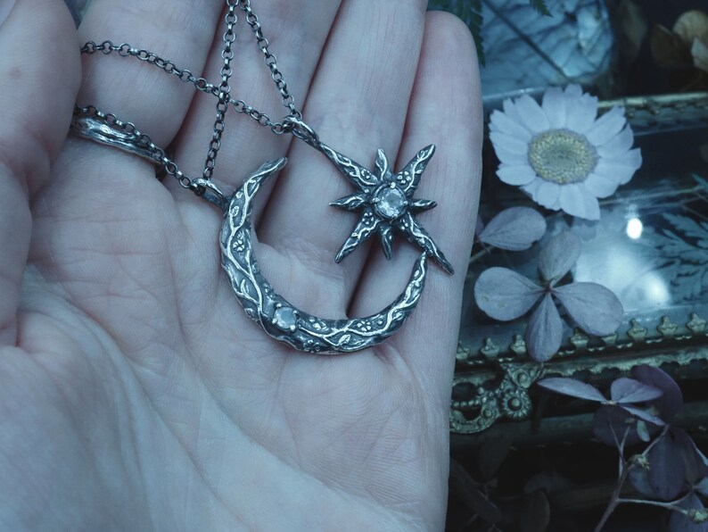 May include: A silver necklace with a crescent moon and star pendant. The pendant is detailed with intricate floral designs and features a clear gemstone in the center of the star.