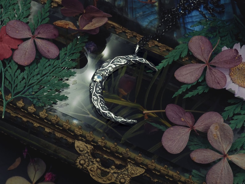 May include: A silver crescent moon pendant necklace with a delicate floral design and a blue gemstone. The necklace is displayed in a vintage-style box with dried flowers.