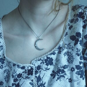 May include: A silver crescent moon pendant necklace on a silver chain. The necklace is worn by a person with a floral patterned blouse.