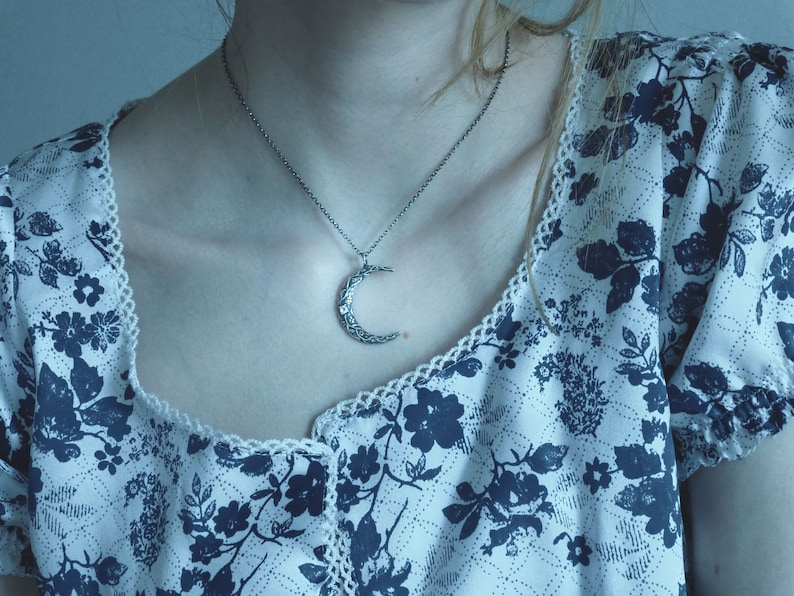 May include: A silver crescent moon pendant necklace on a silver chain. The necklace is worn by a person with a floral patterned blouse.