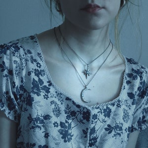 May include: A woman wearing a white blouse with a blue floral pattern and two silver necklaces. One necklace features a crescent moon pendant and the other features a star pendant.