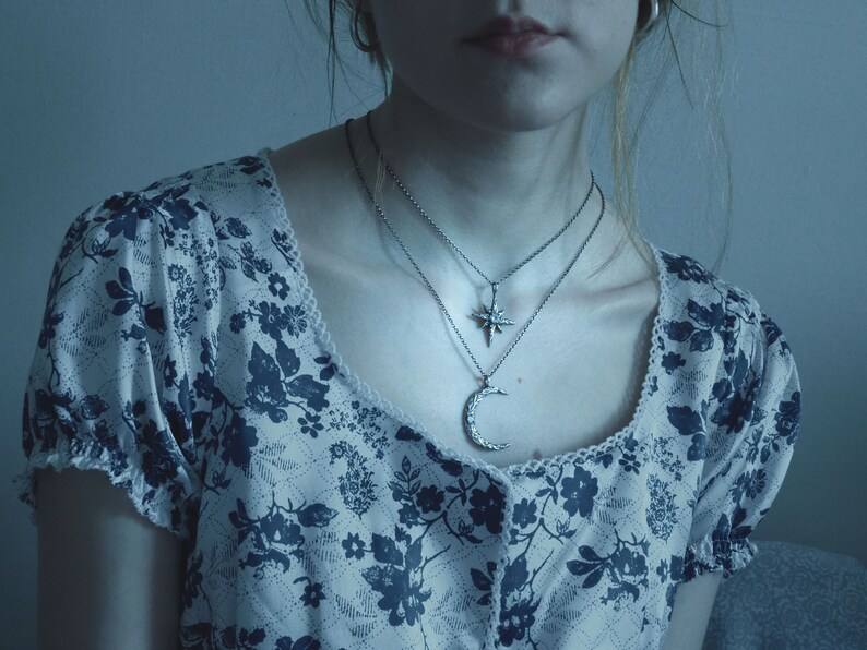 May include: A woman wearing a white blouse with a blue floral pattern and two silver necklaces. One necklace features a crescent moon pendant and the other features a star pendant.