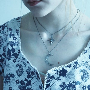 May include: A woman wearing a white blouse with a blue floral pattern and two silver necklaces. One necklace features a crescent moon pendant and the other features a star pendant.