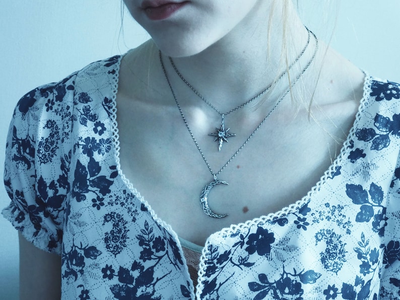 May include: A woman wearing a white blouse with a blue floral pattern and two silver necklaces. One necklace features a crescent moon pendant and the other features a star pendant.