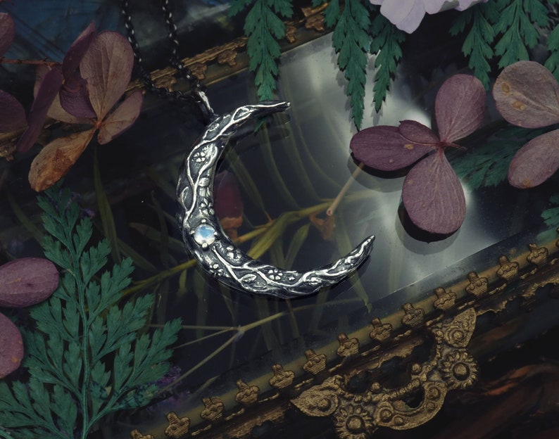 May include: A silver crescent moon pendant necklace with a delicate floral design and a clear gemstone in the center. The necklace is displayed on a dark background with dried flowers.