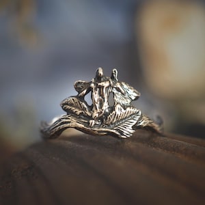 May include: A bronze-toned ring featuring a sculpted design of two figures with outstretched arms, surrounded by stylized leaves. The ring has a nature-inspired theme, with intricate details and a vintage aesthetic.
