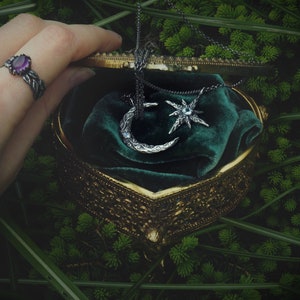May include: A vintage-style jewelry box with a green velvet interior, containing a silver crescent moon and star pendant necklace. The box is open and a hand is reaching in to retrieve the necklace.