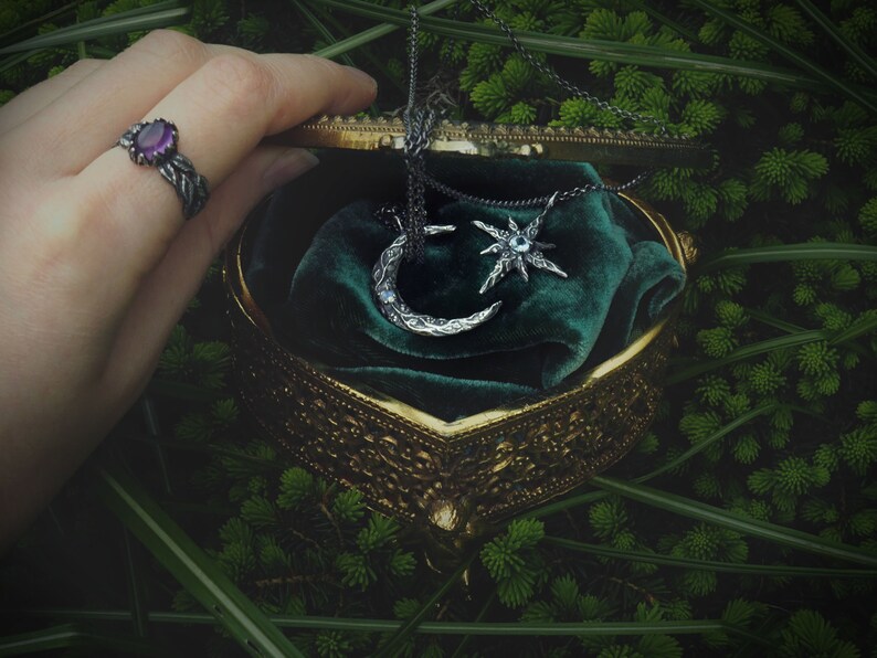May include: A vintage-style jewelry box with a green velvet interior, containing a silver crescent moon and star pendant necklace. The box is open and a hand is reaching in to retrieve the necklace.