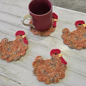 May include: A red mug sits on a crocheted chicken-shaped coaster. Three additional coasters are visible. The coasters are made of orange, yellow, and brown yarn, with red combs and yellow beaks. The coasters are on a light-colored wooden surface.