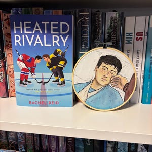 May include: A book titled "Heated Rivalry" with a hockey-themed cover, next to a round embroidery hoop art piece. The book is by Rachel Reid. The embroidery depicts a person in a hospital bed.