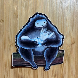 May include: A black and blue sticker featuring a large, furry creature with a smiling face holding a smaller, glowing creature in its arms. The larger creature is sitting on a branch.