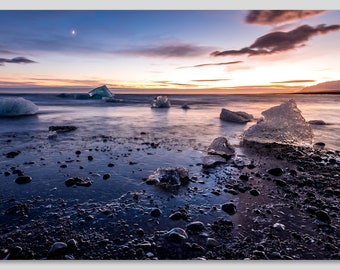 May include: A scenic landscape photograph of a beach at dusk. Icebergs are scattered along the shoreline, reflecting the colors of the sunset. The sky transitions from blue to orange, with a crescent moon visible.