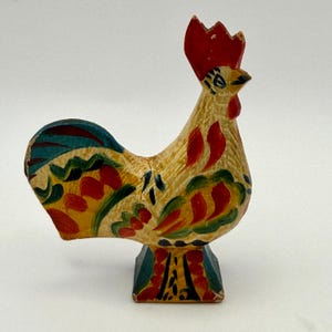 May include: A hand-painted wooden rooster figurine featuring a bright red comb and wattle. The body is decorated with yellow, green, blue, and red painted patterns. The rooster stands on a square base with matching colorful painted details.