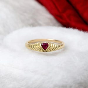 May include: A gold ring with a heart-shaped, deep red gemstone at the centre. The ring's band features a ribbed design, and the heart-shaped stone is set in a gold bezel. The ring is displayed on a white, textured surface.