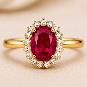 May include: A gold ring featuring a large, oval-cut deep red gemstone at the center, surrounded by a halo of small, round, clear white stones. The band is smooth and polished gold, creating a classic and elegant appearance for this jewelry piece.