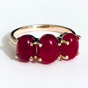 May include: A gold ring with three oval, deep red gemstones. The gemstones are set in a row across the top of the ring, held by gold prongs. The ring band is a simple, polished gold. The gemstones appear to be approximately 0.5 cm in diameter.