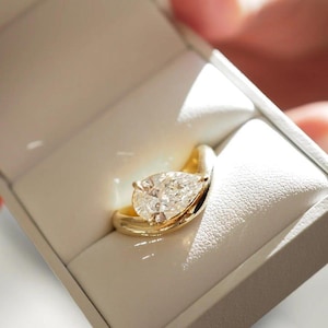Puede incluir: Un anillo de oro con un diamante en forma de pera, presentado en una caja de joyería blanca. La banda del anillo se curva elegantemente alrededor del diamante. El diamante es claro y brilla, reflejando la luz.