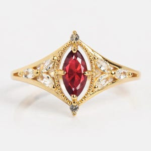 Op de afbeelding: Een gouden ring met een marquise-geslepen, dieprode edelsteen in het midden. De ring is versierd met kleinere, heldere edelstenen en gedetailleerde gouden details. Het ontwerp is symmetrisch en elegant, geschikt voor speciale gelegenheden.