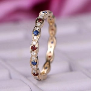 Może przedstawiać: Złoty pierścionek z powtarzającym się wzorem czerwonych, niebieskich i białych kamieni szlachetnych. Pierścionek posiada szereg owalnych sekcji, z których każda zawiera mały kamień szlachetny. Kamienie szlachetne są ułożone w linii wokół pierścionka.