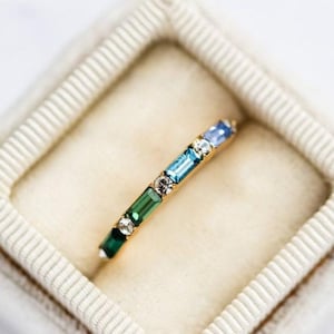 May include: A gold band ring with a row of colorful gemstones. The gemstones are green, blue, and light blue. The ring is in a white velvet ring box.