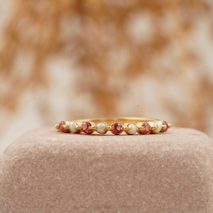 Peut inclure: Une bague en or ornée de pierres précieuses rouges et blanches alternées. La bague est posée sur une pierre brun clair, avec un arrière-plan flou de plantes séchées. La bague présente de petites pierres rondes.