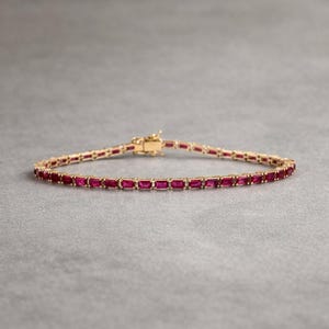 May include: A gold bracelet with a series of rectangular, deep red gemstones. The bracelet is clasped and lies on a gray surface. The gemstones are evenly spaced and set in a gold band.