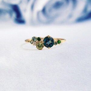 Può includere: Un anello d'oro con un gruppo di pietre preziose blu, verdi e bianche. La pietra più grande è una pietra preziosa rotonda blu.