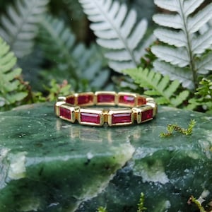 May include: A gold ring with rectangular, deep red gemstones set around the band. The ring is displayed on a green stone surface, with a backdrop of green foliage.