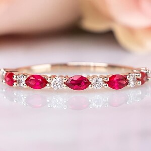 Könnte beinhalten: Ein zarter Ring mit abwechselnd markiseförmigen roten Edelsteinen und runden, klaren Edelsteinen, die in einem Goldband gefasst sind. Die Edelsteine sind gleichmäßig um den Ring angeordnet. Der Ring wird auf einer reflektierenden Oberfläche präsentiert.