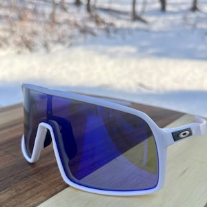 May include: White-framed sunglasses with a large, wraparound design and blue mirrored lenses. The sunglasses have a modern, sporty aesthetic, with a sleek, curved frame and a small logo on the temple.