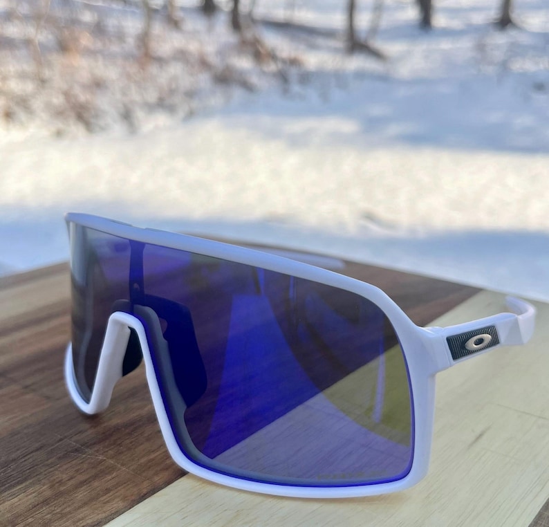 May include: White-framed sunglasses with a large, wraparound design and blue mirrored lenses. The sunglasses have a modern, sporty aesthetic, with a sleek, curved frame and a small logo on the temple.