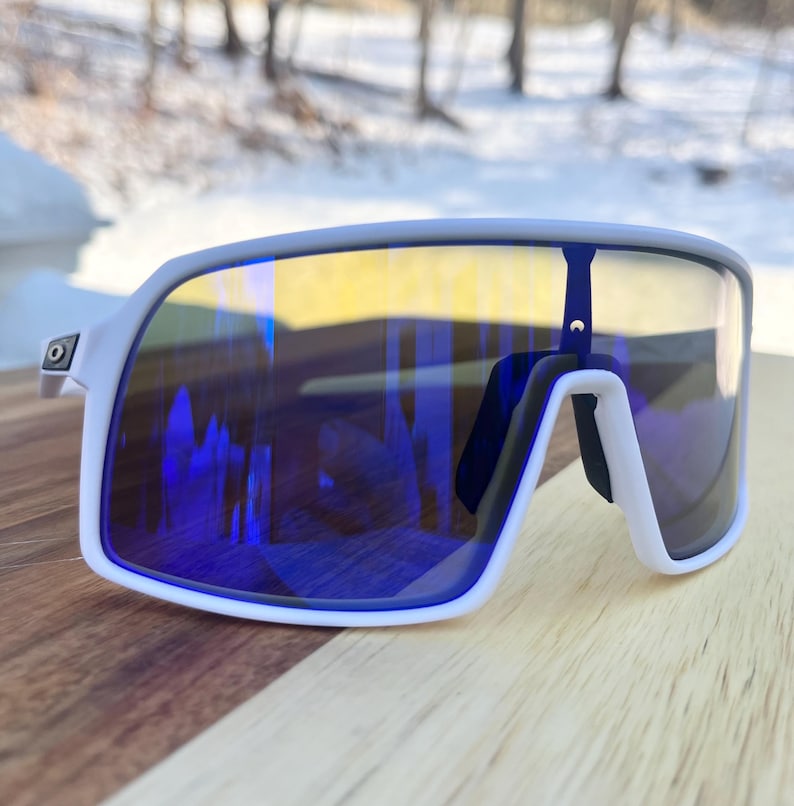 May include: White sports sunglasses with blue and yellow mirrored lenses. The sunglasses have a thick white frame and a black nose bridge. The lenses reflect the surrounding environment, creating a vibrant and dynamic appearance.
