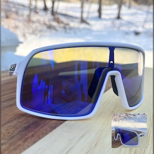 May include: White-framed sports sunglasses with blue and yellow mirrored lenses. The sunglasses have a modern, angular design and a small, secondary image of the sunglasses in the bottom right corner. The background shows a snowy landscape.