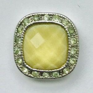 May include: A square-shaped, yellow, faceted glass button with a silver metal frame and a border of light green rhinestones.
