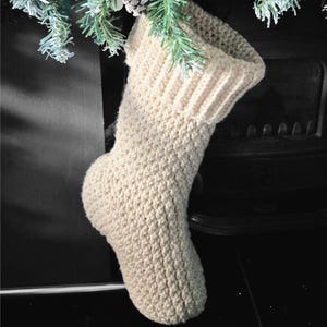 May include: A cream-coloured, crocheted Christmas stocking hangs from a decorative evergreen bough. The stocking has a ribbed cuff and a textured body. The text "Crochet Pattern" and "Instant Digital Download" are displayed.