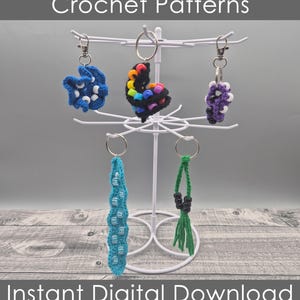 Puede incluir: Cuatro patrones de llavero de ganchillo que se muestran en un soporte de metal blanco. Los llaveros están hechos con cuentas de colores y hilo de lana en colores azul, blanco, morado, verde, negro y arcoíris. Descarga digital instantánea.