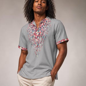 May include: A light gray short-sleeved shirt featuring a V-neckline and a geometric pattern in shades of pink, blue, and white. The pattern forms a large V-shape on the chest and accents the sleeve cuffs. The shirt is paired with beige trousers.