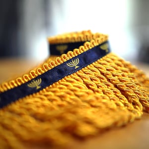 May include: Close-up of a roll of yellow fringe trim with a navy blue ribbon. The ribbon is adorned with gold menorah symbols. The fringe is thick and braided, creating a textured appearance. The trim is likely used for decorative purposes.