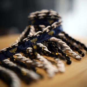 May include: A close-up of a blue ribbon with gold menorah designs, with black and silver twisted cords hanging from the bottom.