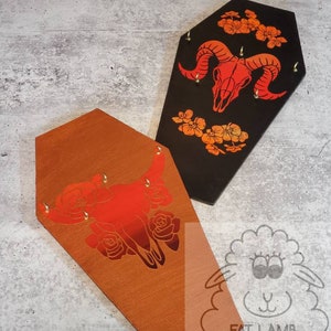 May include: Two wooden coffin-shaped earring holders with a red and orange skull design and gold accents. The holder on the left is brown and the holder on the right is black.